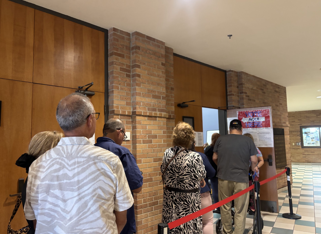 People standing in line to vote. A red sign on a door that reads “Vote Here” and an arrow pointing into a room with the word “Aqui” on it.